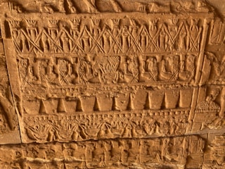 Detailed carvings on a stone surface display rows of intricate figures, symbols, and patterns. The relief features human-like figures and various geometric shapes, all executed with precise craftsmanship. The stone has an aged, tactile texture and a warm, sandstone hue.