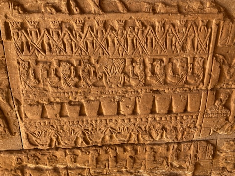 Detailed carvings on a stone surface display rows of intricate figures, symbols, and patterns. The relief features human-like figures and various geometric shapes, all executed with precise craftsmanship. The stone has an aged, tactile texture and a warm, sandstone hue.