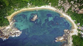 Aerial view of a crescent-shaped beach lined with lush green vegetation. The coastline features rocky outcrops and clear turquoise water, with a small pier extending into the bay. The surrounding area is dotted with a few small structures and a winding path.