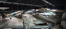 Multiple military aircraft are displayed in a large, indoor museum hangar. The planes vary in size, design, and era, with some having propellers and others jet engines. The hangar has high ceilings and is well-lit, with information boards and barriers around each exhibit. The surface is concrete, and the ambient lighting gives a somewhat somber and historical feel.