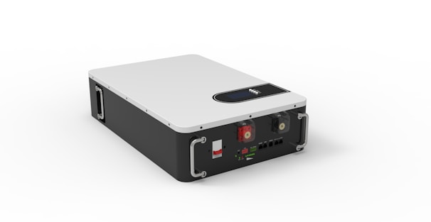 A rectangular device with a sleek design features a white top surface and black sides. It includes metal handles on the front corners for carrying. The front panel has various connectors and switches, including two red and green rotary knobs, and multiple input/output ports.