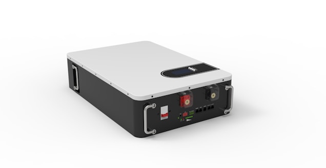 A rectangular device with a sleek design features a white top surface and black sides. It includes metal handles on the front corners for carrying. The front panel has various connectors and switches, including two red and green rotary knobs, and multiple input/output ports.