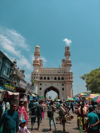A bustling street scene in front of a historic monument with multiple arches and minarets. Numerous people are walking and shopping at small market stalls lining the sides of the street. The sky is clear with some clouds, adding a vibrant blue background to the scene.