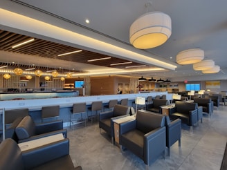 A modern lounge area with gray leather seating and sleek tables. The space is designed with contemporary lighting fixtures, including large pendant lights and warm-toned accent lights. The ceiling features wooden slats, adding texture and visual interest. The bar area is prominent, with stools and a backlit counter. The room is spacious and well-lit, creating a comfortable and inviting atmosphere.