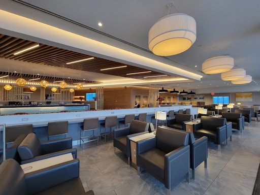 A modern lounge area with gray leather seating and sleek tables. The space is designed with contemporary lighting fixtures, including large pendant lights and warm-toned accent lights. The ceiling features wooden slats, adding texture and visual interest. The bar area is prominent, with stools and a backlit counter. The room is spacious and well-lit, creating a comfortable and inviting atmosphere.