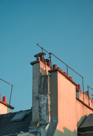 A rooftop with a series of red chimneys aligned along the edge of a building, bathed in warm sunlight. The structure features old weathered walls and metal railings, creating an architectural silhouette against a clear blue sky.