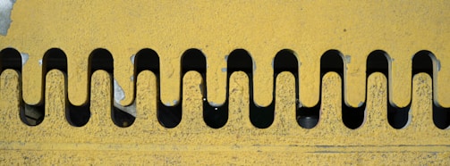 A close-up of a textured yellow metal surface featuring a pattern of symmetrical wavy cutouts. The cutouts reveal a dark background, creating a contrasting effect. The metal surface shows signs of wear and some peeling paint, indicating exposure to the elements.