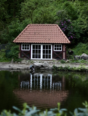 A small, rustic house with a terracotta tiled roof is situated in a lush, green forested area. The house has large windows and a reflective surface of water in front creating a clear reflection. Surrounding the house are dense trees and plants, with some purple flowers adding contrast to the greenery.