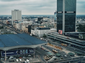 An urban landscape featuring a busy city intersection with several yellow trams passing through. High-rise buildings, including one with prominent Samsung branding, dominate the skyline. A large building with a roof sign reads 'WARSZAWA CENTRALNA' indicating a central location. The weather appears overcast, adding a muted tone to the city scene.