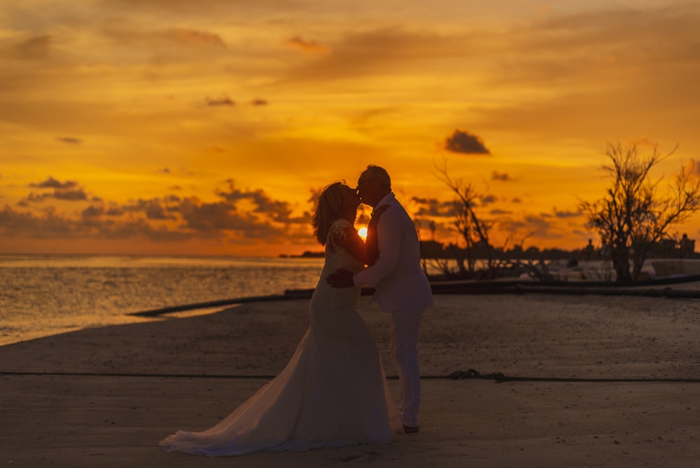 a bride and groom kissing on the beach at sunset