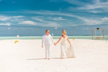 A couple is walking hand in hand on a pristine white sandy beach under a vibrant blue sky. The woman is wearing a long white dress, resembling a wedding gown, while the man is dressed in a light-colored outfit. The background features turquoise ocean waters and the horizon. A wooden swing is visible on the right side of the image.