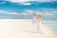 A couple stands on a sandy beach, wearing wedding attire with the groom in a light suit and the bride in a flowing white gown. They embrace each other affectionately against a backdrop of a bright blue sky and calm ocean waters.