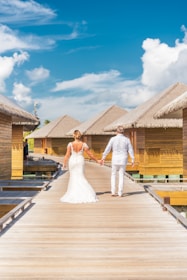 A couple is walking hand in hand along a wooden pathway, surrounded by tropical overwater bungalows with thatched roofs. The woman is wearing a wedding dress, and the man is dressed in a light-colored outfit. The sky is clear with some white clouds, creating a serene and romantic atmosphere.