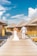 A couple is walking hand in hand along a wooden pathway, surrounded by tropical overwater bungalows with thatched roofs. The woman is wearing a wedding dress, and the man is dressed in a light-colored outfit. The sky is clear with some white clouds, creating a serene and romantic atmosphere.
