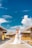 A couple dressed in wedding attire stands on a wooden boardwalk surrounded by overwater bungalows. The woman is wearing a white wedding gown and the man is in a white suit. They are embracing and smiling under a bright blue sky with scattered white clouds.