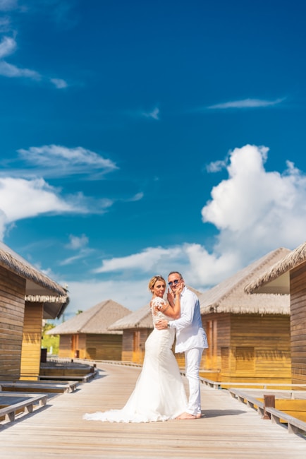 A couple dressed in wedding attire stands on a wooden boardwalk surrounded by overwater bungalows. The woman is wearing a white wedding gown and the man is in a white suit. They are embracing and smiling under a bright blue sky with scattered white clouds.