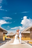 A couple in wedding attire embraces on a wooden walkway with thatched-roof bungalows on either side under a bright blue sky with scattered clouds.