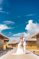 A couple in wedding attire embraces on a wooden walkway with thatched-roof bungalows on either side under a bright blue sky with scattered clouds.