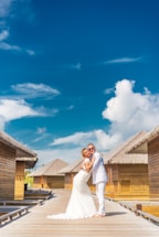 A couple in wedding attire embraces on a wooden walkway with thatched-roof bungalows on either side under a bright blue sky with scattered clouds.