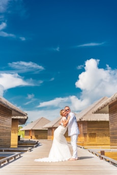 A couple in wedding attire embraces on a wooden walkway with thatched-roof bungalows on either side under a bright blue sky with scattered clouds.