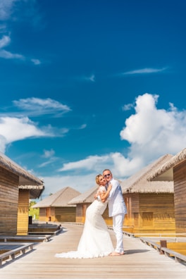A couple in wedding attire embraces on a wooden walkway with thatched-roof bungalows on either side under a bright blue sky with scattered clouds.