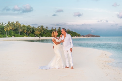 A couple stands on a serene beach with clear turquoise waters and a backdrop of lush palm trees. They are dressed in elegant white attire, embracing affectionately. The sky has a hint of clouds with a soft pastel hue.
