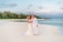 A couple stands on a serene beach with clear turquoise waters and a backdrop of lush palm trees. They are dressed in elegant white attire, embracing affectionately. The sky has a hint of clouds with a soft pastel hue.