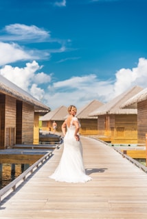 A bride in a white gown stands on a wooden boardwalk beside overwater bungalows. The sky is bright blue with scattered clouds, creating a picturesque tropical setting.