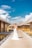 A bride in a white gown stands on a wooden boardwalk beside overwater bungalows. The sky is bright blue with scattered clouds, creating a picturesque tropical setting.