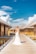 A bride in a white gown stands on a wooden boardwalk beside overwater bungalows. The sky is bright blue with scattered clouds, creating a picturesque tropical setting.