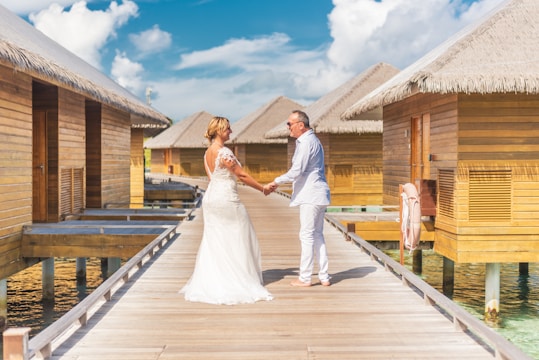 A couple stands hand in hand on a wooden walkway lined with thatched-roof huts over clear, blue water. The woman wears a white wedding dress, and the man is dressed in a light-colored suit. The sky is clear with a few fluffy clouds, creating a serene and romantic setting.