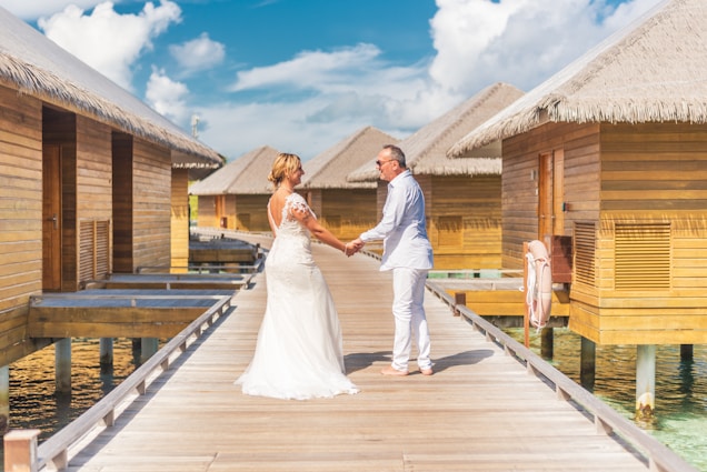 A couple stands hand in hand on a wooden walkway lined with thatched-roof huts over clear, blue water. The woman wears a white wedding dress, and the man is dressed in a light-colored suit. The sky is clear with a few fluffy clouds, creating a serene and romantic setting.