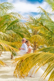 A couple is embracing, surrounded by lush palm trees on a sunny beach. The woman is wearing a white wedding dress and the man is dressed in a white shirt. They appear relaxed and affectionate amidst a tropical setting.