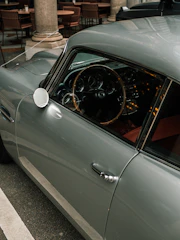 A classic car with a glossy exterior and vintage design is parked next to a row of empty outdoor tables and chairs, creating a serene and nostalgic atmosphere.
