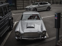 A silver classic car is parked in a city street, flanked by a modern SUV and another vehicle. The scene includes urban elements like a parking meter and city architecture in the background.