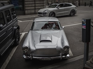 A silver classic car is parked in a city street, flanked by a modern SUV and another vehicle. The scene includes urban elements like a parking meter and city architecture in the background.