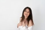 A young woman with long dark hair stands against a plain white background, smiling warmly. She is wearing a white outfit with an off-the-shoulder style and has her hands gently placed near her chest.