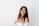 A young woman with long dark hair stands against a plain white background, smiling warmly. She is wearing a white outfit with an off-the-shoulder style and has her hands gently placed near her chest.
