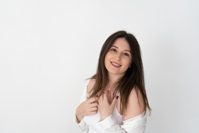 A young woman with long dark hair stands against a plain white background, smiling warmly. She is wearing a white outfit with an off-the-shoulder style and has her hands gently placed near her chest.