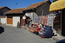 A street with a shop displaying colorful rugs and textiles hanging on the wall. Various artisanal goods are arranged on stands outside the shop. A person is seated on a chair nearby, possibly overseeing the merchandise. Traditional tiled roofs and a clear blue sky are seen in the background.