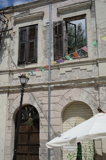 A weathered building facade with two upper-level windows showcasing wooden shutters, one of which is open. Below, there are two archways, one with a dark wooden door and the other with a partially visible painted scene on a roll-up door. Colorful triangular pennant flags are strung across the front, and a black street lamp stands to the left. An outdoor umbrella is partially visible at the bottom right.