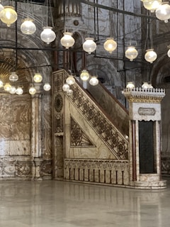 An intricately designed marble staircase leads up with an ornate railing, set within a richly decorated interior featuring stylish patterns. Hanging spherical lamps provide warm illumination, enhancing the elegant historical architecture with decorative archways and columns.