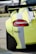 A close-up view of the rear section of a sports car, showcasing its sleek design and rear spoiler. The car is painted in a bright yellow color with white stripes, and features a large rear tire. The Mobil 1 logo is prominently displayed on the side.