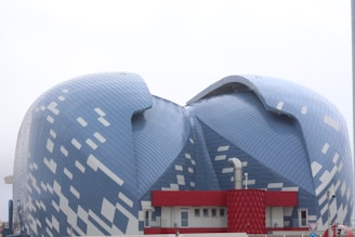 A modern, uniquely designed building with a blue, patterned facade resembling overlapping scales or tiles. It features a dynamic, organic shape with large, curved surfaces. The lower section includes a red structure with geometric details and windows, adding contrast to the blue exterior.
