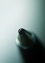 A macro shot of the tip of a ballpoint pen, highlighting its metallic point and the reflection of light on its surface. The background appears to be a soft gradient of green.