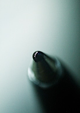 A macro shot of the tip of a ballpoint pen, highlighting its metallic point and the reflection of light on its surface. The background appears to be a soft gradient of green.