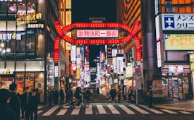 A bustling urban street scene filled with neon lights and colorful signs. The crowded street is lined with shops and buildings, and a large illuminated sign in Japanese script arches over the entrance. Pedestrians cross a zebra crossing and a person on a bicycle is visible. The nighttime setting highlights the vibrant city atmosphere.