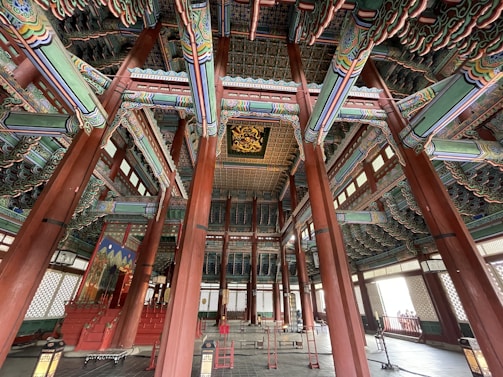 An intricately decorated interior space with colorful patterns on wooden columns and beams. The architecture features rich detailing on the ceiling and a harmonious blend of colors. The room has a spacious, symmetrical layout, with traditional Asian design elements throughout.