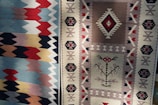 Two intricately patterned woven textiles with distinctive geometric designs and rich colors are displayed side by side. The left textile features a series of zigzag patterns in shades of blue, red, and cream. The right textile displays a more complex arrangement with diamond motifs and smaller geometric shapes in beige, brown, and red tones.