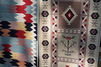 Two intricately patterned woven textiles with distinctive geometric designs and rich colors are displayed side by side. The left textile features a series of zigzag patterns in shades of blue, red, and cream. The right textile displays a more complex arrangement with diamond motifs and smaller geometric shapes in beige, brown, and red tones.